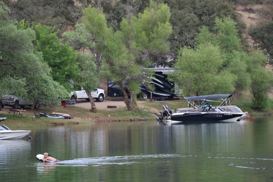A wake boat and paddler on calm green water in front of camp