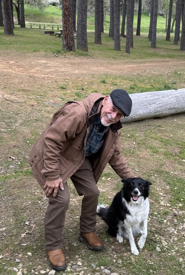 A man in a tan jacket petting a black-and-white border collie in a pine campground