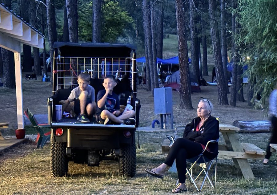 Two children sitting in the back of a utility cart at dusk while an adult sits nearby in a camp chair