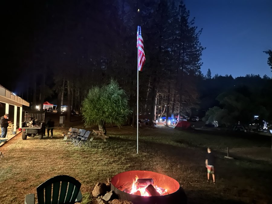 A campfire burning in a fire ring at night with a lit flagpole and a dog crossing the lawn