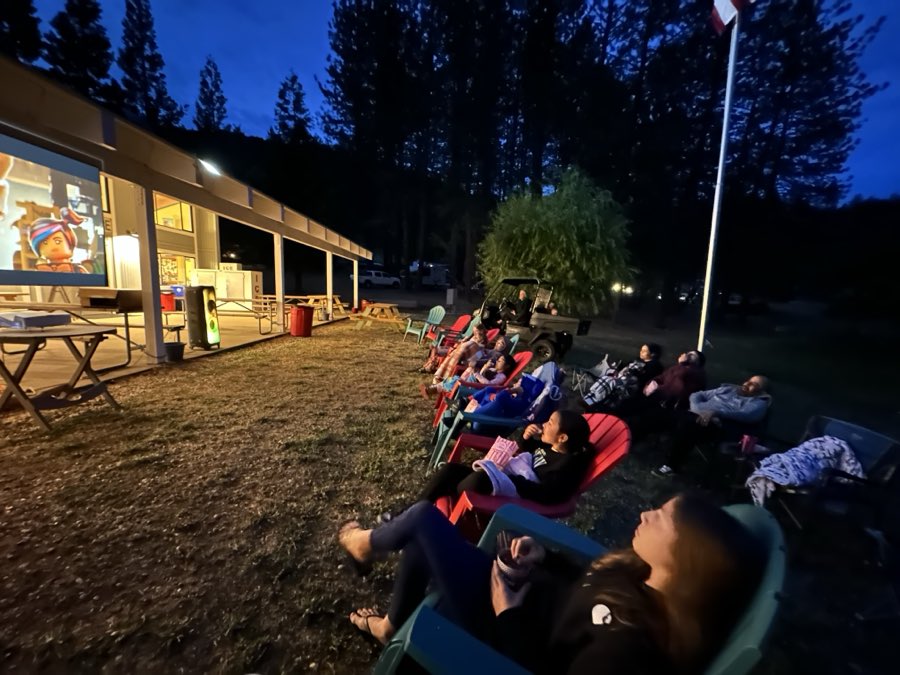 A crowd in camp chairs on a lawn at dusk watching an outdoor movie at the lodge